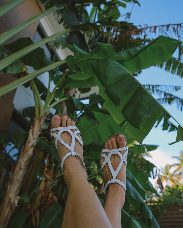 Image shows a models feet wearing the Dorith Sandal in white. The feet are up in the air. There are some banana trees against a blue sky in the background which give a tropical vibe to the shot.