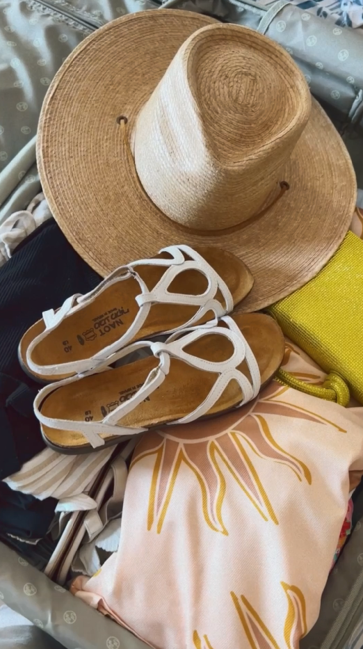 Picture shows a suitcase open with a stylish sun hat, the Naot Sandal Dorith in white sitting on top of the neatly piled suitcase. Othe contents include a swimmin costime towell and slippers.